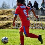 A girl in a red jersey playing soccer outdoors on grass with spectators in background.