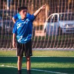 A teenage soccer player in blue jersey directing teammates during a game on a sunny day.